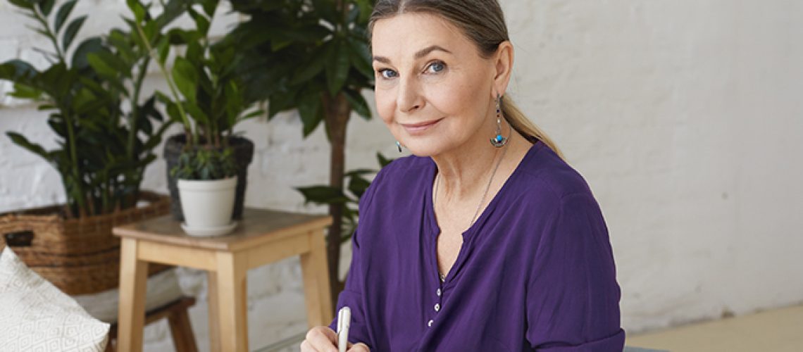 Portrait of busy modern 50 yeard old middle aged woman in violet shirt writing in copybook, making plans, looking at camera with positive friendly smile, sitting on chair, surrounded with green plants